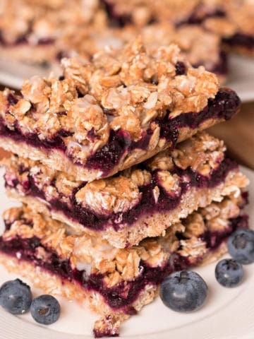 Three Blueberry Oat Bars stacked on a small plate Three Blueberry Oat Bars with lemon glaze stacked on a small plate. More bars in the background.