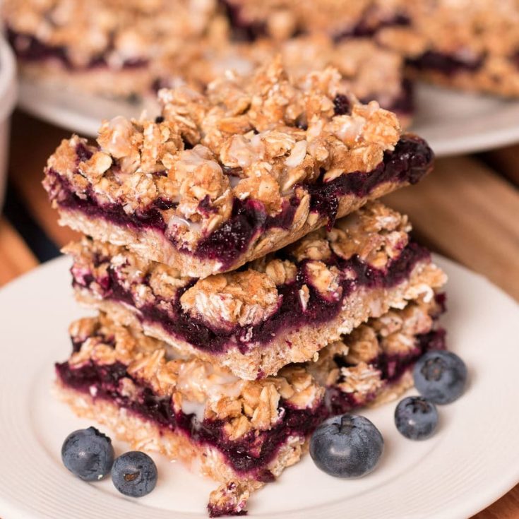 Three Blueberry Oat Bars stacked on a small plate Three Blueberry Oat Bars with lemon glaze stacked on a small plate. More bars in the background.