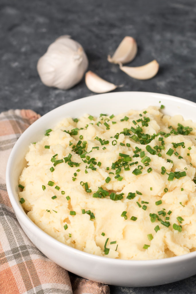Bowl of Vegan Roasted Garlic Mashed Potatoes with chives sprinkled on top and garlic in the background