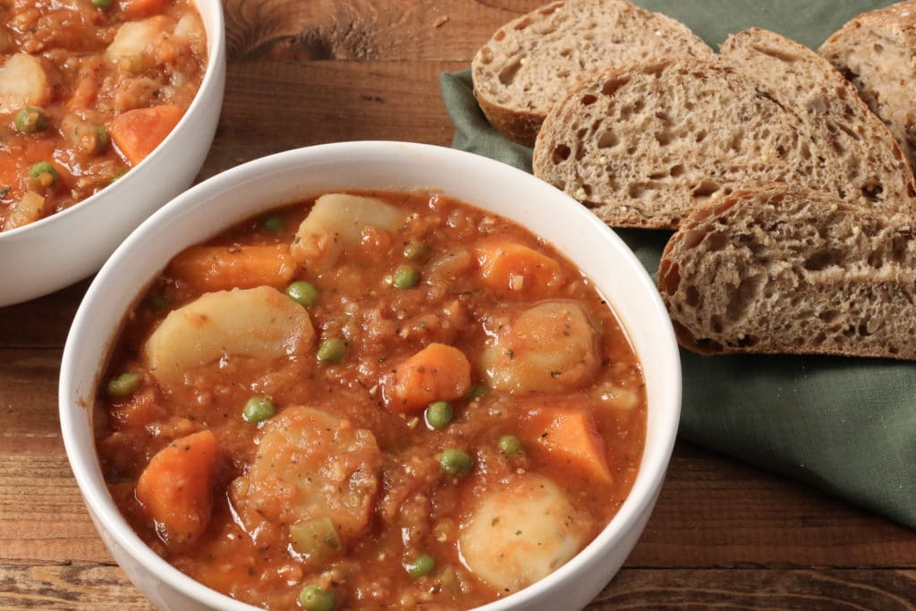 Bowl of Instant Pot Vegetable Stew in a white bowl, sitting on a wooden surface, with another bowl of stew and sliced multi-grain bread in the background
