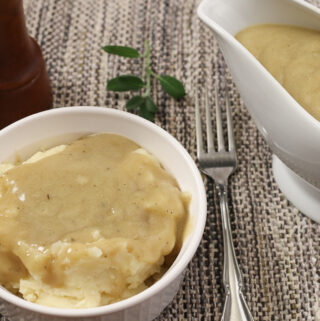 Horizontal image of bowl of mashed potatoes and gravy boat with Easy Vegan Golden Gravy sitting on a placemat with fresh sage, a fork, and pepper mill in background