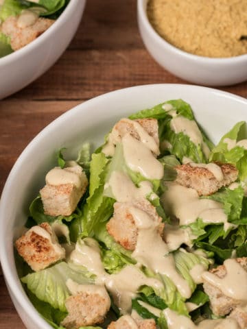Photo of two bowls of Vegan Caesar Salad on wooden board, with jar of Caesar dressing and bowl of nutritional yeast in background