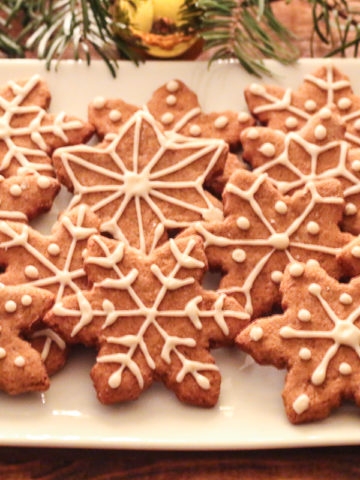 White plate of Oil-Free Vegan Gingerbread cookies, cut into snowflake shapes. Fir branches and red and gold Christmas ornaments surround the plate.