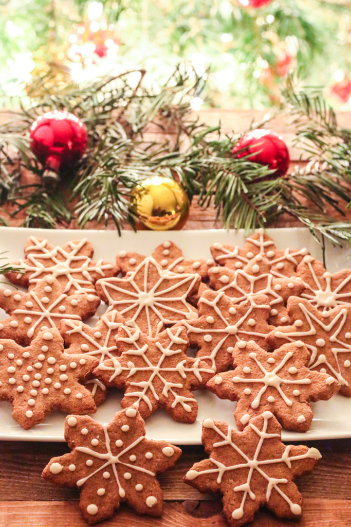 White plate of Oil-Free Vegan Gingerbread cookies, cut into snowflake shapes. Fir branches and red and gold Christmas ornaments surround the plate. Lit Christmas tree branches in background.