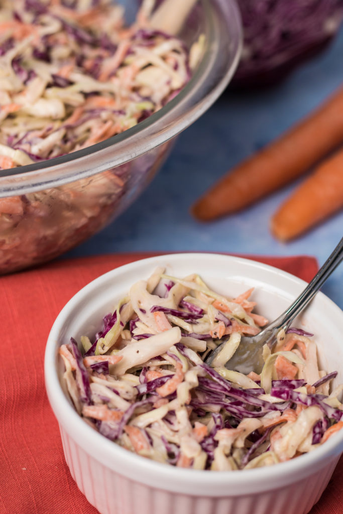 Small white bowl of vegan coleslaw sitting on red napkin, alongside larger bowl of coleslaw, with carrots and cabbage in background