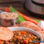 Bowl of lentil soup with spoon and slice of bread in the bowl; more bread, carrots, celery, garlic, and lentils in the background, Pinterest title above image