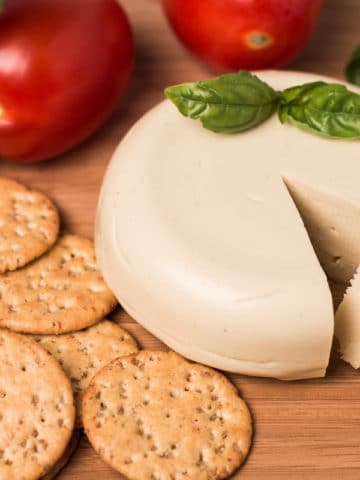 Wheel of vegan cheese with wedge cut out of it, tomatoes, basil, and crackers, background