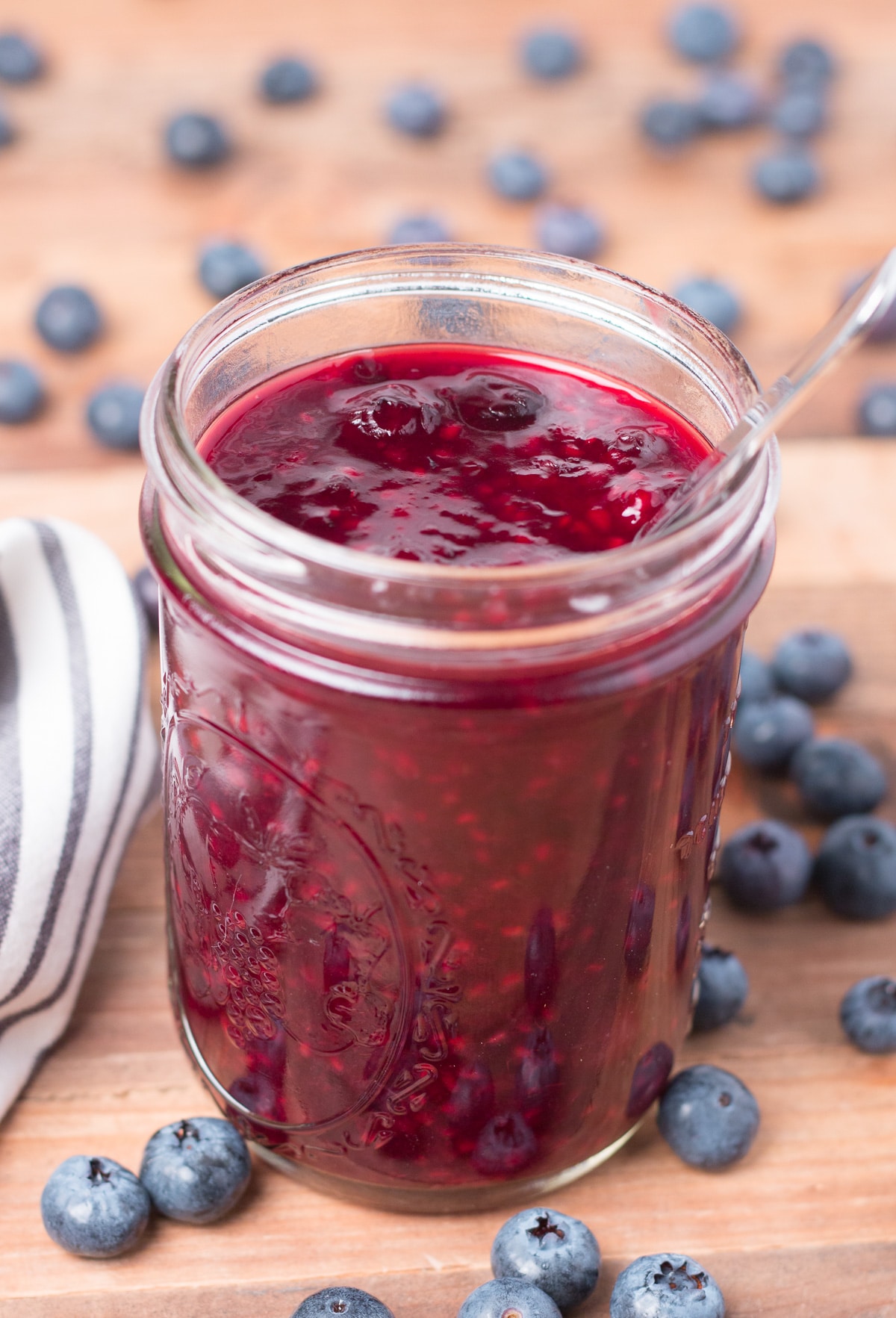 Jar of berry sauce on a wooden surface with striped napkin beside it and blueberries scattered around it