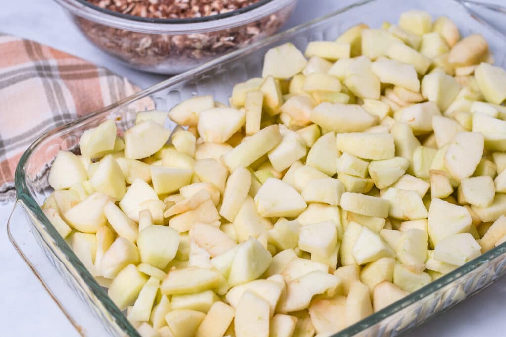 Glass baking pan of apple slices with napkin and bowl of oatmeal toppin in background