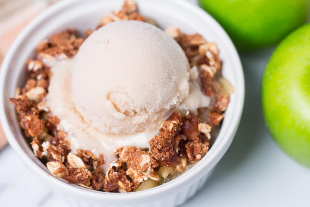 Small white bowl of apple crisp with a scoop of vanilla ice cream, two green apples beside it