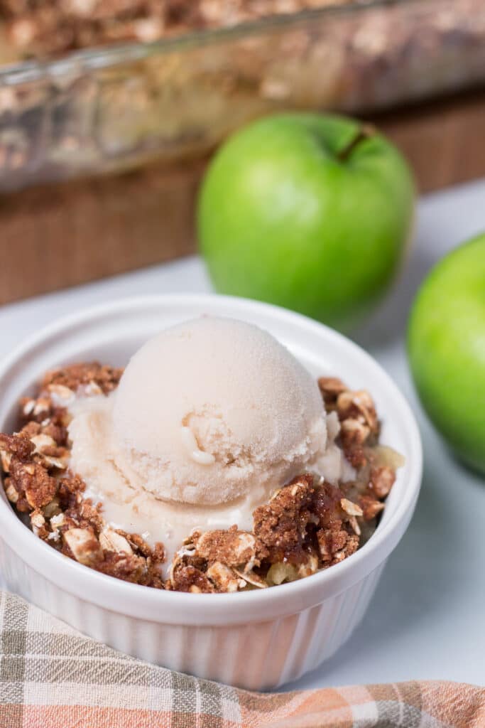 Small white bowl of apple crisp with a scoop of vanilla ice cream on top, apples and pan of apple crisp in the background
