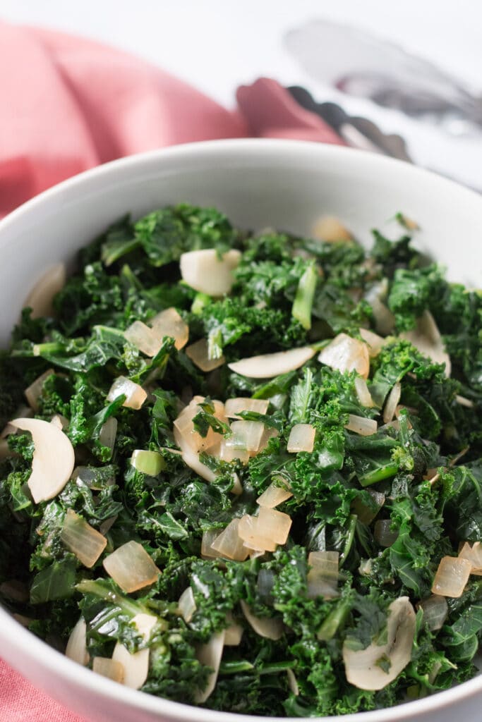 White bowl of kale with garlic and onions, with pink napkin beside bowl and metal tongs in background