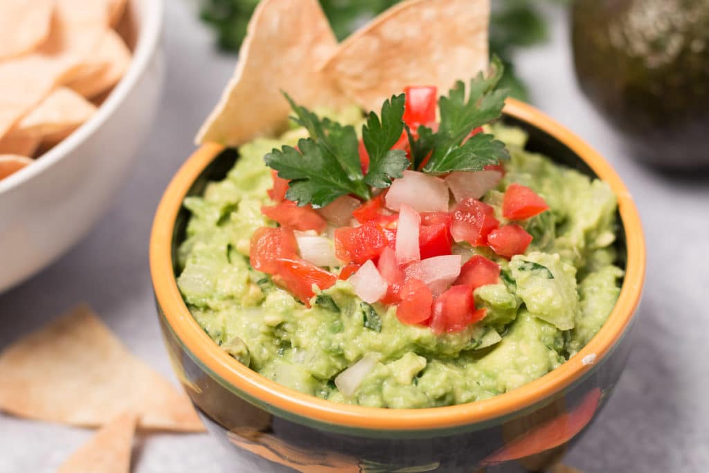 Bowl of guacamole with cilantro, avocado, and bowl of chips in background