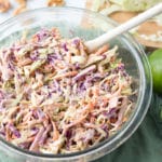 Bowl of coleslaw with limes beside it and cutting board with cabbage in the background