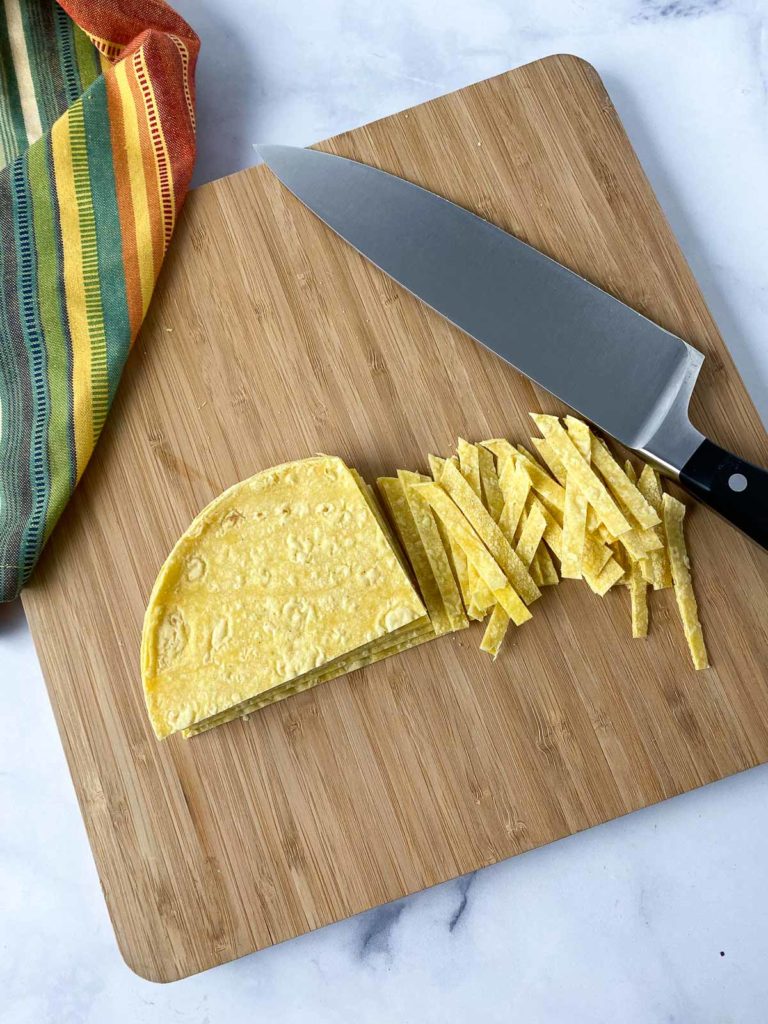 Corn tortillas partially cut into strips and a knife on a wooden cutting board