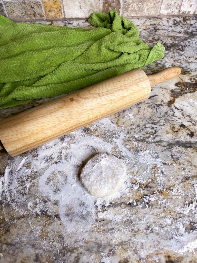 Flattened ball of dough on floured countertop with rolling pin and covered dough in background