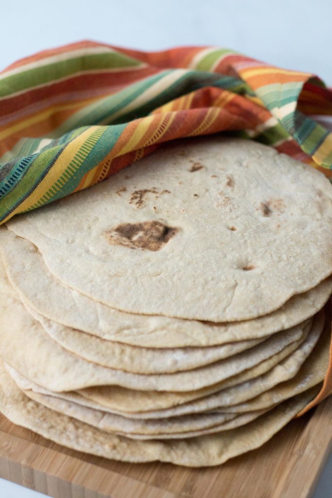 Stack of whole wheat tortillas ona wooden cutting board partially covered with striped towell