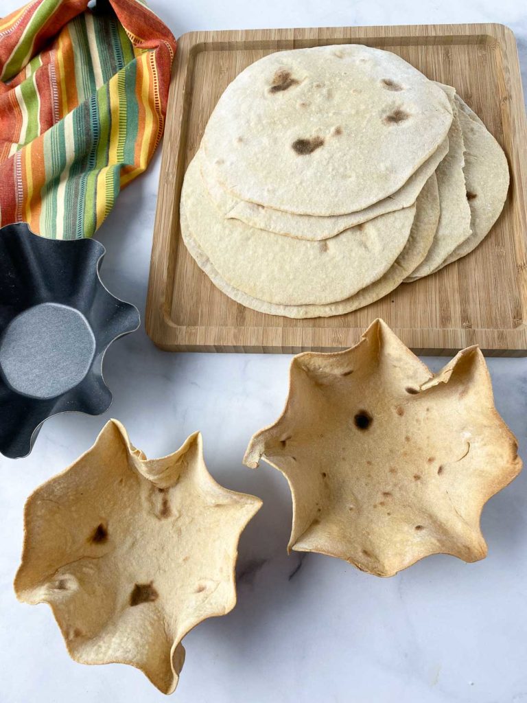 Two baked tortilla bowls in front of stack of tortillas on wooden cutting board