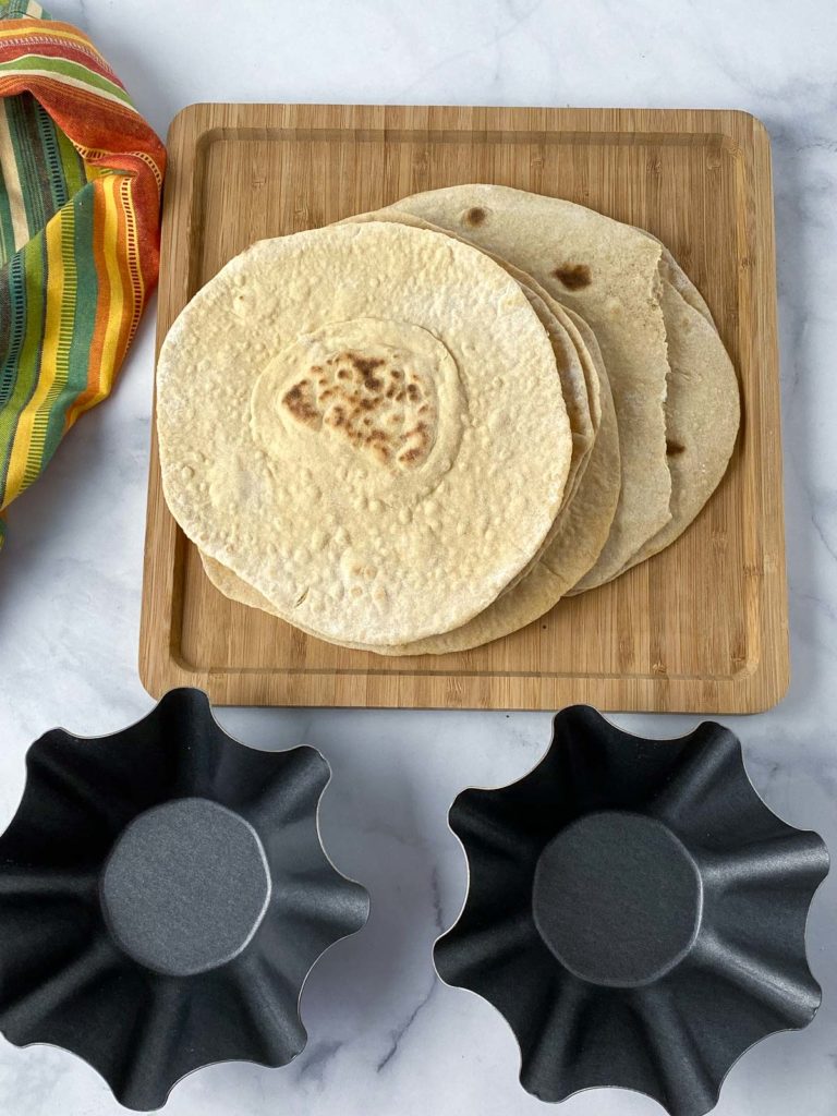 Two tortilla bowl molds in front of stack of tortillas on a wooden cutting board