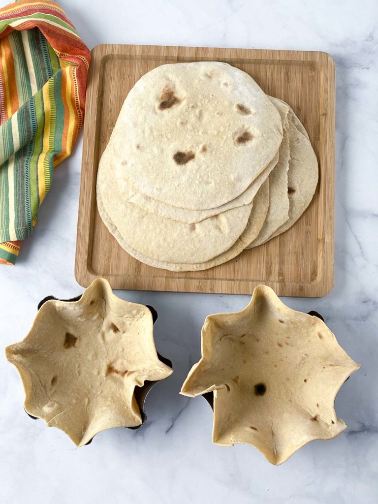 Two tortillas in tortilla bowl molds in frong of stack of tortillas on wooden cutting board