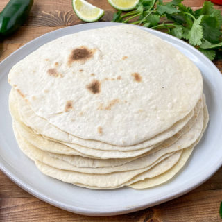 Stack of fat free tortillas on a plate with jalapenos, lime, tomatoes, and cilantro around them