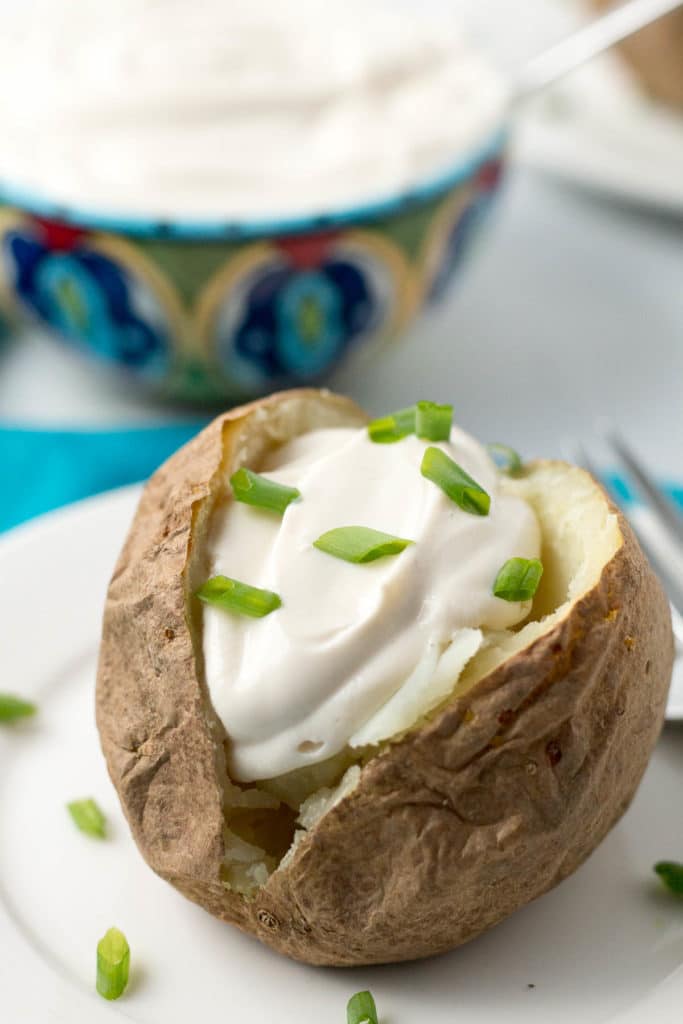 Baked potato topped with sour cream and green onions with bowl of sour cream in background