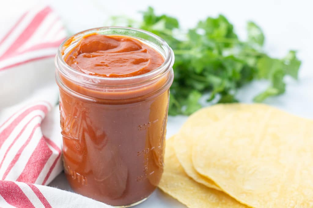 Jar of red sauce next to striped napkin and corn tortillas with cilantro in background.