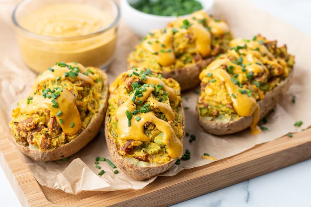 Four twice baked potatoes on a cutting board with a bowl of cheese sauce and a bowl of chives.
