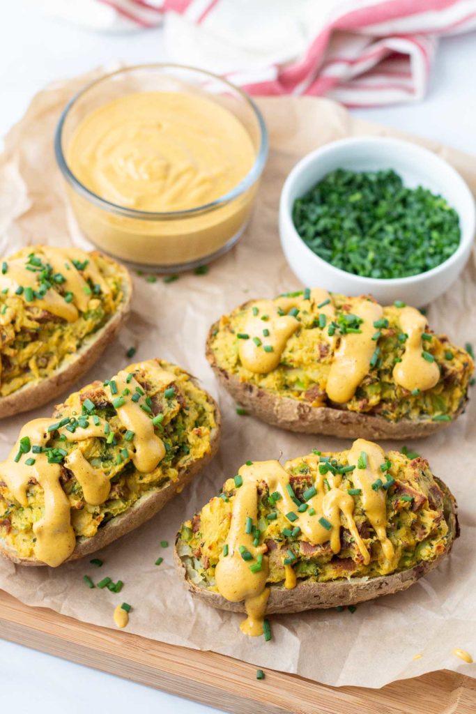 Twice baked potatoes on a cutting board with a bowl of cheese sauce and a bowl of chives.