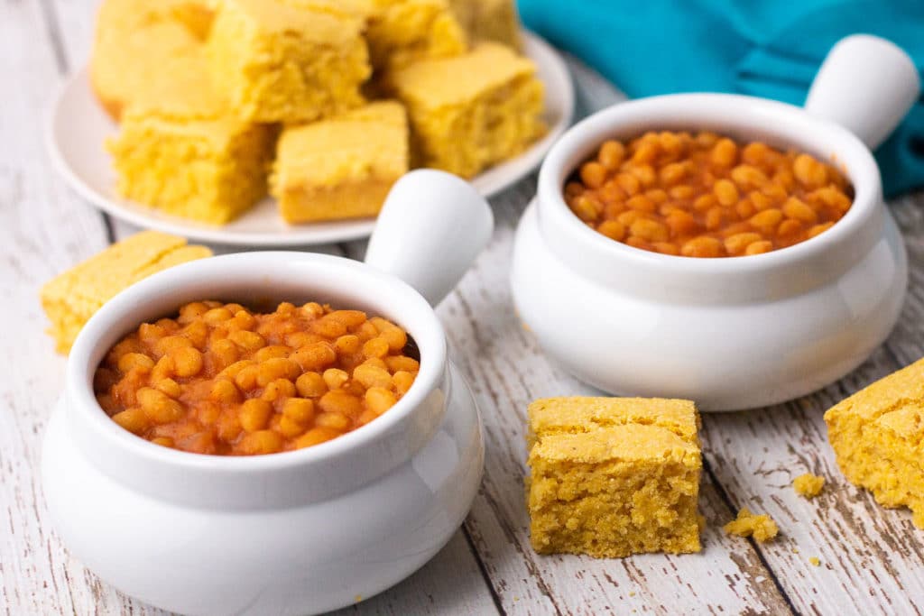 Two bowls of baked beans with plate of cornbread in background.