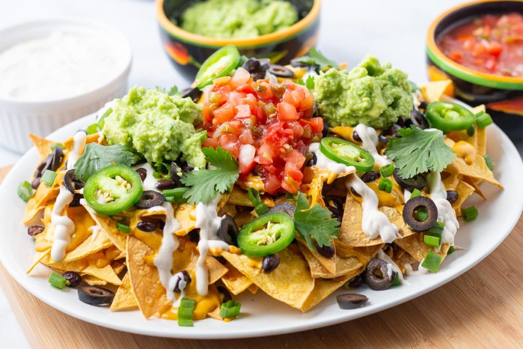 Platter of nachos with bowls of guacamole, salsa, and sour cream in background.