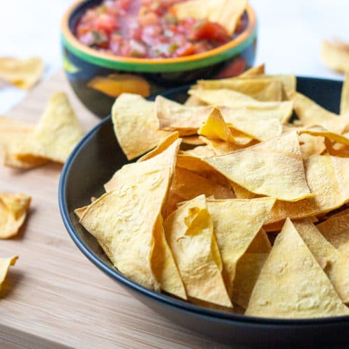 Bowl of tortilla chips with scattered chips and bowl of salsa in the background.
