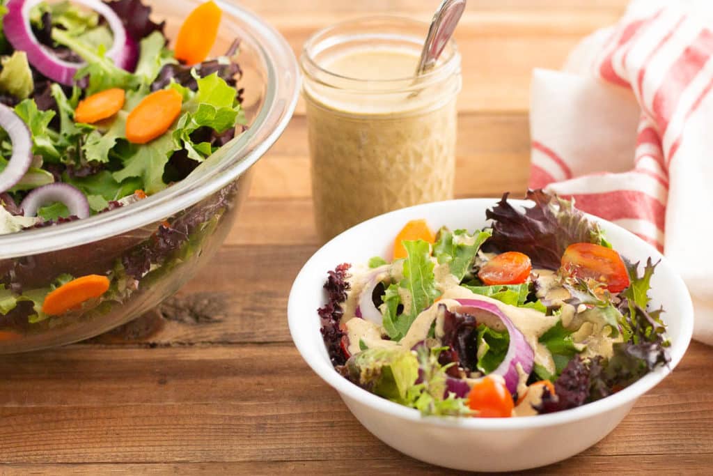 Serving of green salad with jar of salad dressing and large bowl of salad in the background