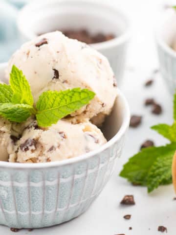 Bowl of vegan mint chocolate chip ice cream with fresh mint leaf garnish, another bowl of ice cream, bowl of chopped chocolate, and ice cream cones in background.