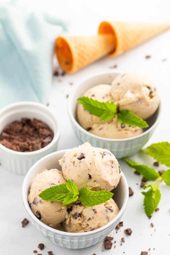 Two bowls of vegan mint chocolate chip ice cream with fresh mint leaf garnish, with bowl of chopped chocolate and ice cream cones in background.