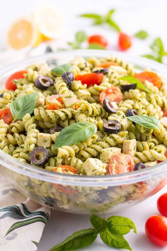 Vegan pesto pasta salad in a glass mixing bowl with basil, tomatoes, and lemon in background.