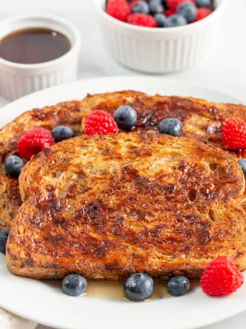 Plate of french toast with syrup and berries, with cup of syrup, bowl of berries, and cup of coffee in background.