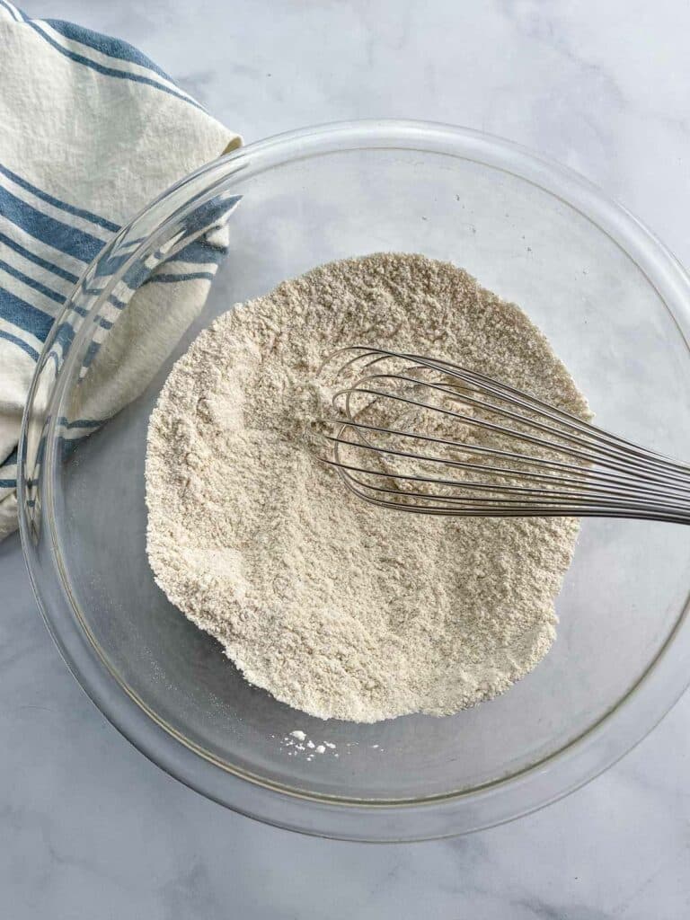 Glass bowl of dry ingredients with a whisk in bowl and cloth napkin on the side.