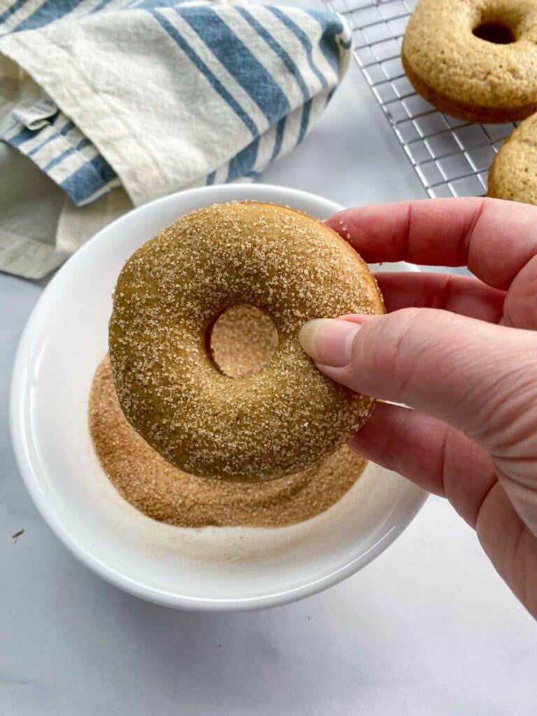 Cinnamon sugar coated donut being held above a bowl of cinnamon sugar with cloth napkin and cooling rack of donuts in background.