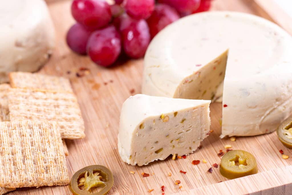 Cheese wheel with wedge cut out of it, with crackers, grapes, and another cheese wheel in background.