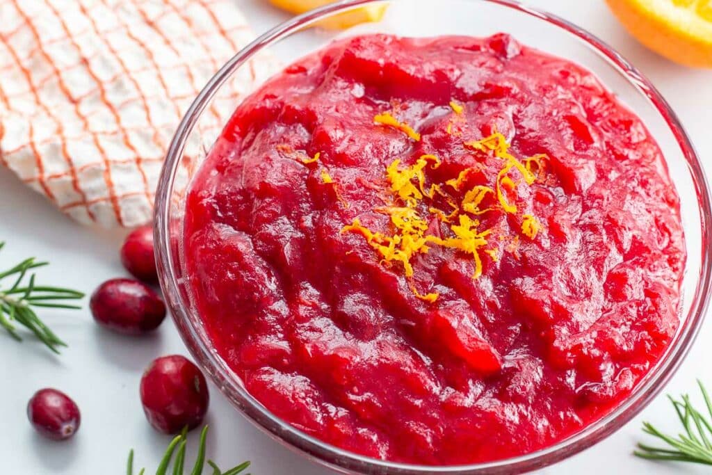 Glass bowl of cranberry sauce garnished with orange zest with fresh cranberries, rosemary, and cloth napkin beside it.