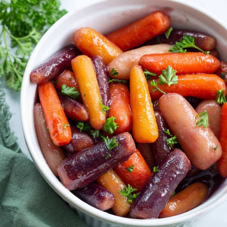 Overhead image of white bowl of multicolored glazed baby carrots next to some parsley and a green napkin.