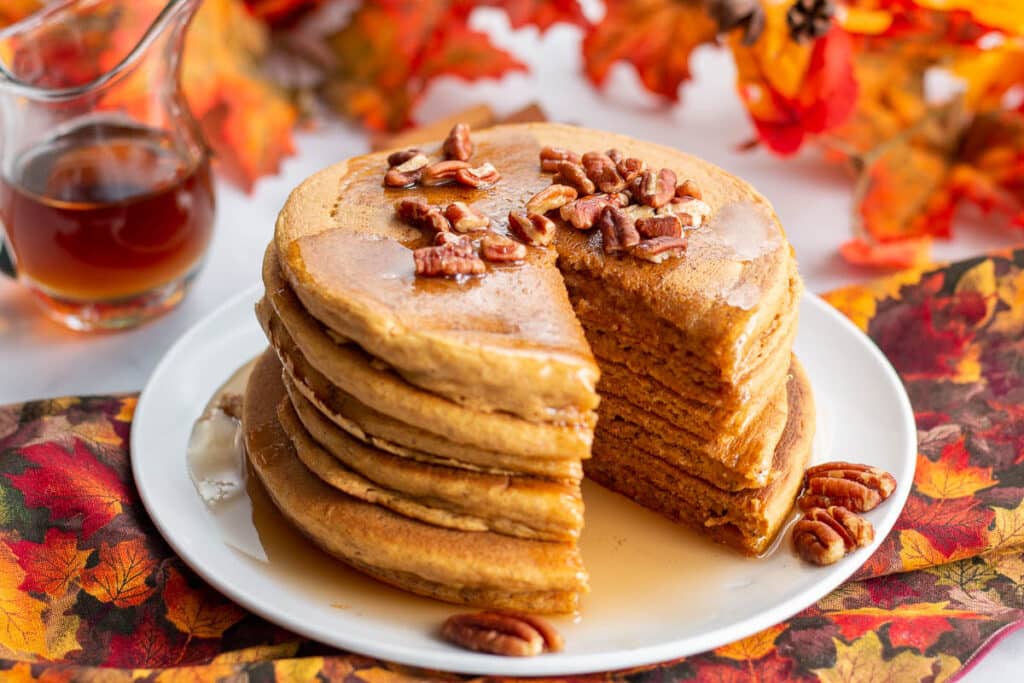 Stack of pecan topped pumpkin pancakes with a wedge cut out of them with syrup pitcher and fall leaves in background.