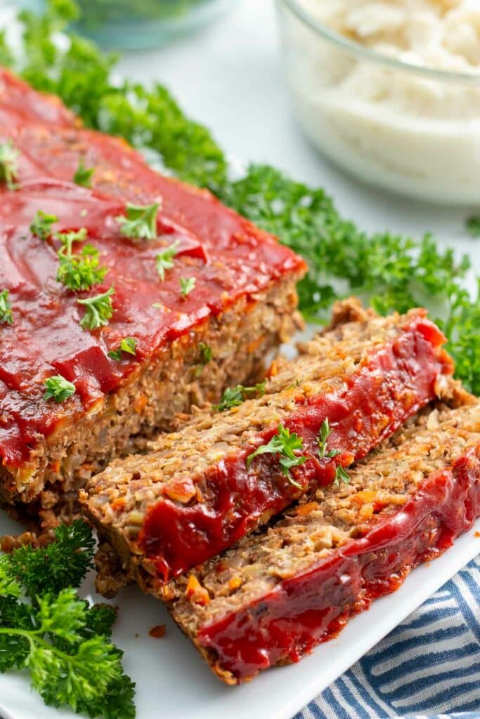 Lentil loaf with 2 slices and parsley on a platter with bowl of mashed potatoes next to it.