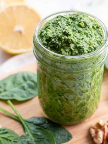 Jar of pesto on wooden cutting board surrounded by spinach leaves, walnuts, and lemon halves.