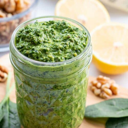 Jar of pesto on wooden cutting board surrounded by spinach leaves, walnuts, and lemon halves.