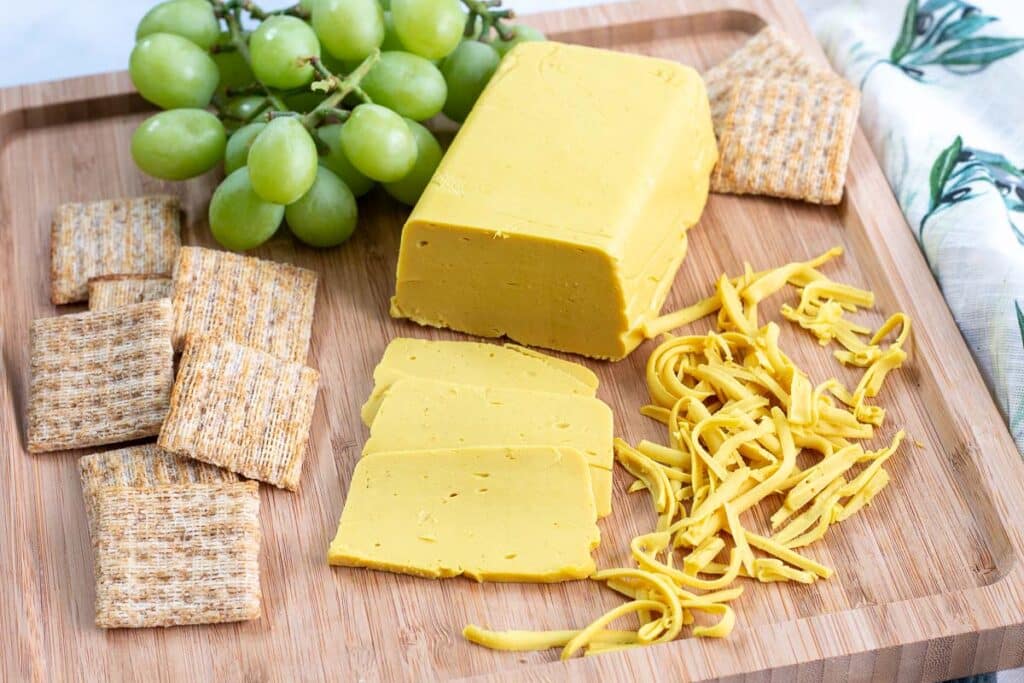 Vegan cheese on wooden cutting board with cheese slices and shreds, crackers, and green grapes.