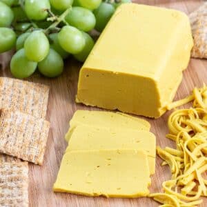 Closeup of vegan cheddar cheese on a cutting board with cheese slices and shreds crackers, and green grapes.