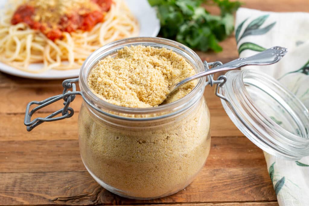 Vegan Parmesan cheese in a glass jar with a plate of spagetti in the background.
