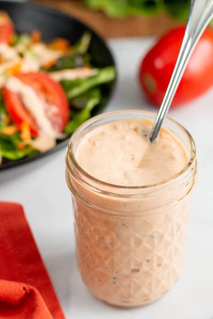 Jar of thousand island dressing with spoon in front of bowl of salad and tomato.